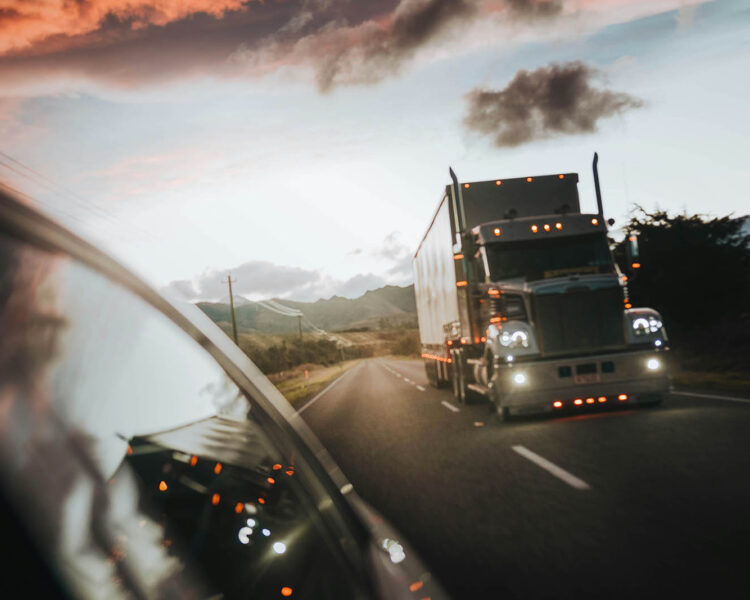 Truck driving at sunset on a scenic highway, with clouds and landscape in the background.