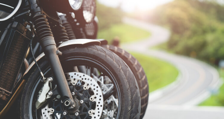Close-up of motorcycle tires on a winding road, with lush greenery and morning sunlight in the background.